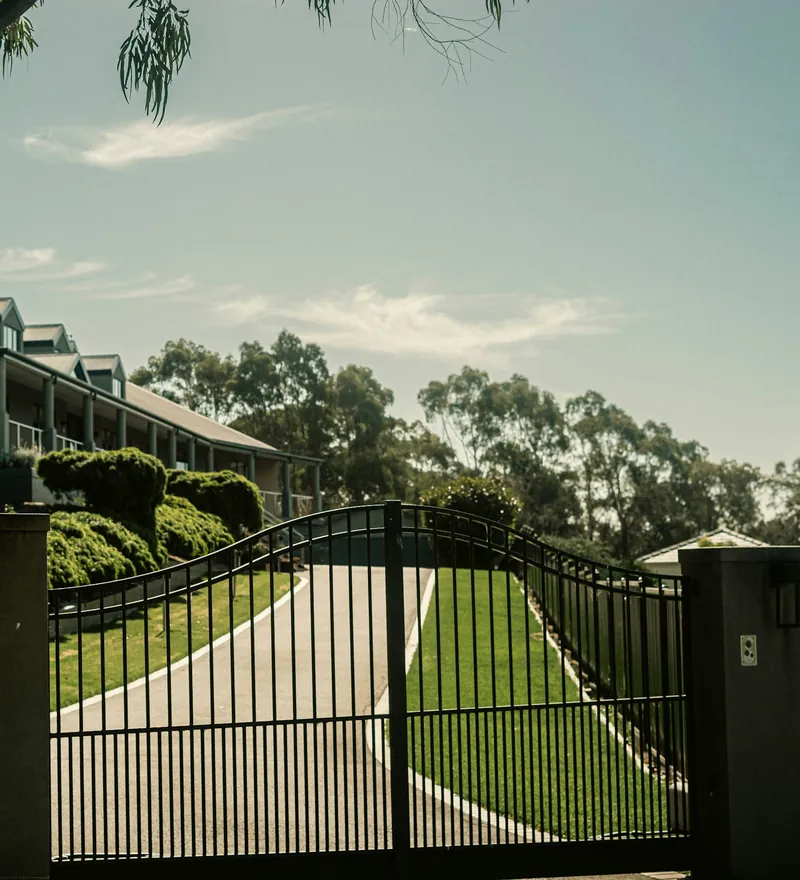 Black aluminum double driveway gate with a keypad post at the foot of a curved driveway lined with trees