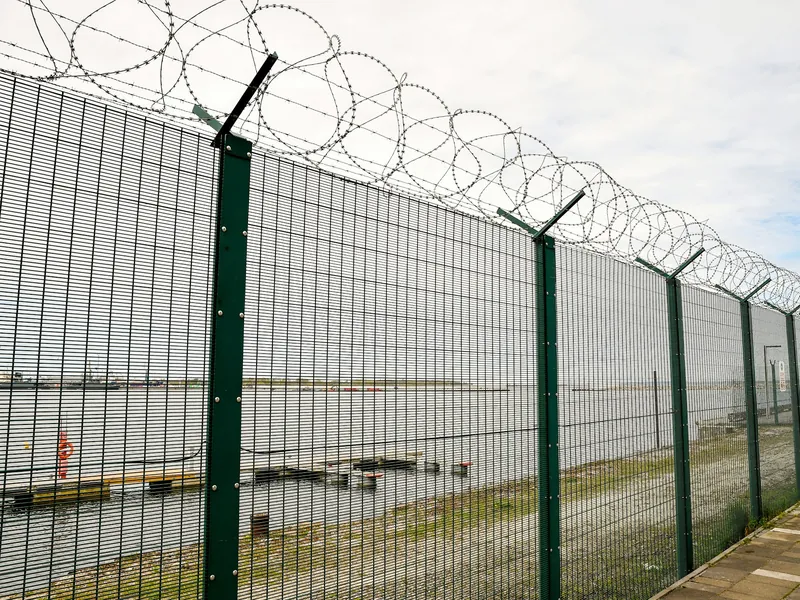 Tall green welded-wire security fence topped with razor wire coils along a paved walkway
