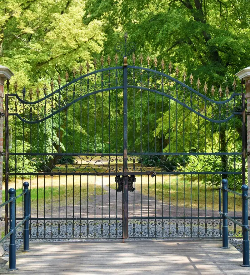 Black wrought iron double driveway gate with scroll details hung between brick pillars, with trees behind