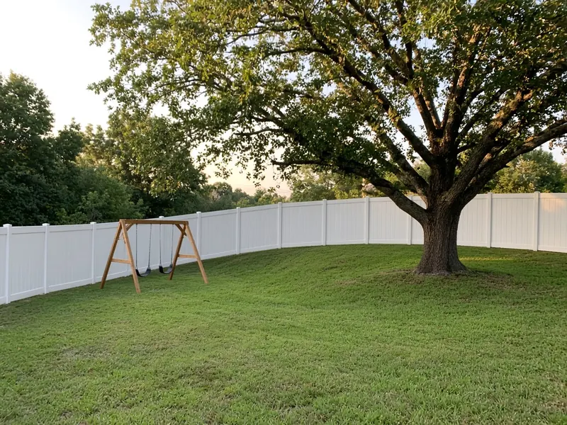 White vinyl privacy fence curving around a backyard with a swing set and shade oak